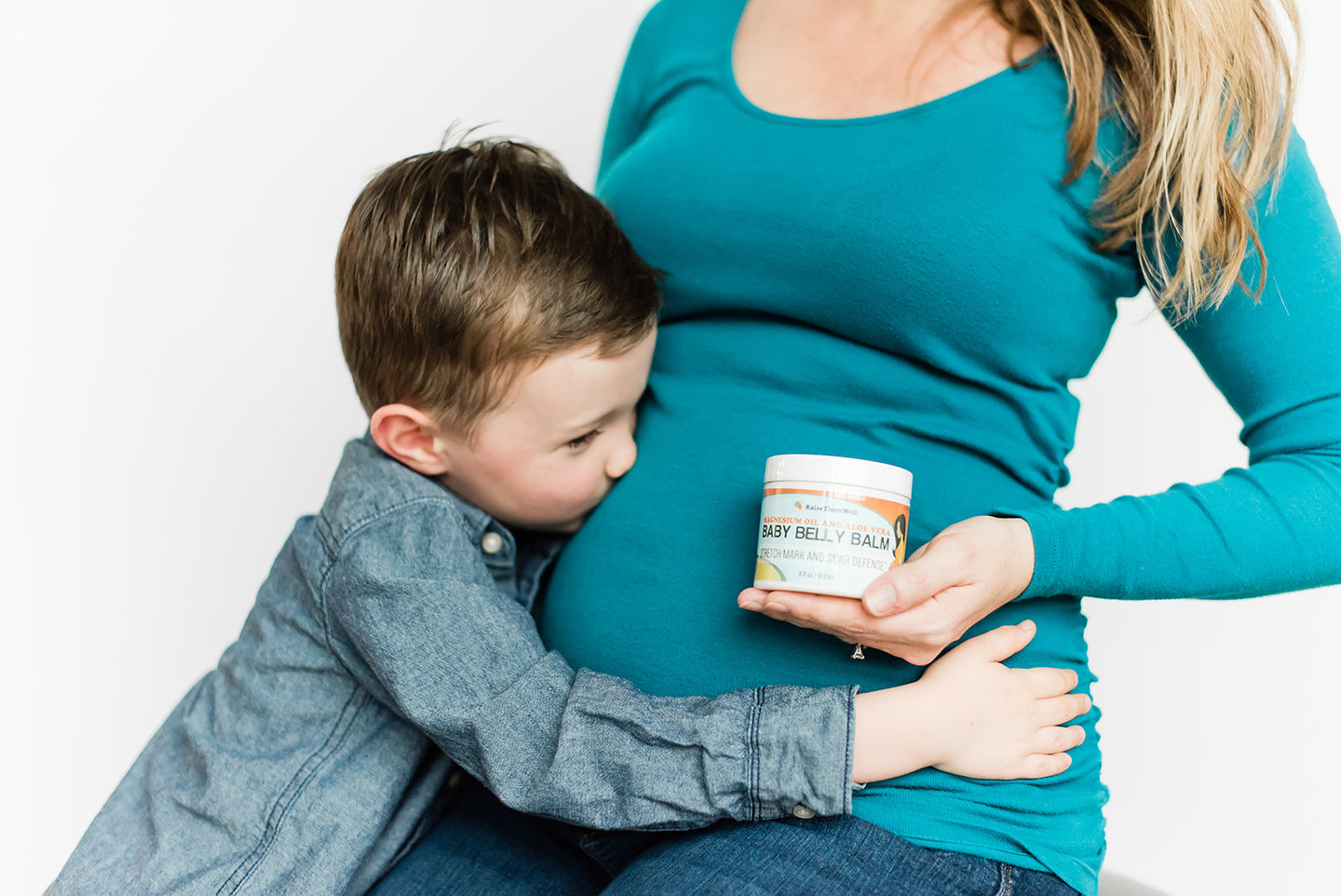 young boy kissing pregnant mom's belly as she prepares to apply baby belly balm stretch mark and scar defense with magnesium oil